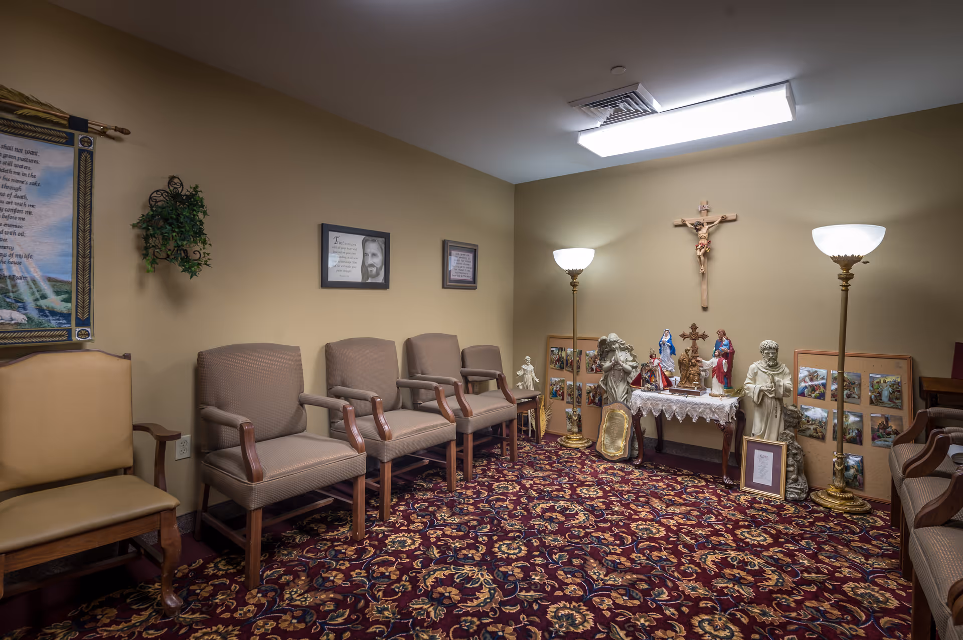 A small room with beige walls and patterned carpet featuring several chairs arranged along the walls. The room contains religious statues, framed pictures, and a crucifix mounted on the wall. Two standing lamps provide lighting.