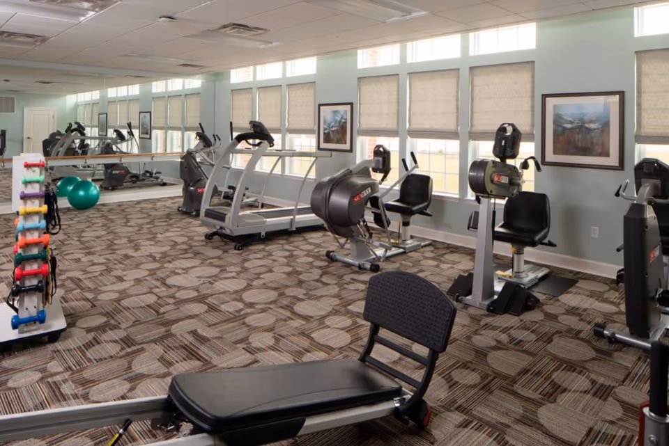 A well-lit fitness room with various exercise equipment including treadmills, stationary bikes, a rowing machine, and a rack of colorful dumbbells. Large windows with blinds allow natural light to fill the room, and a large mirror covers one wall. The floor is carpeted with a patterned design.