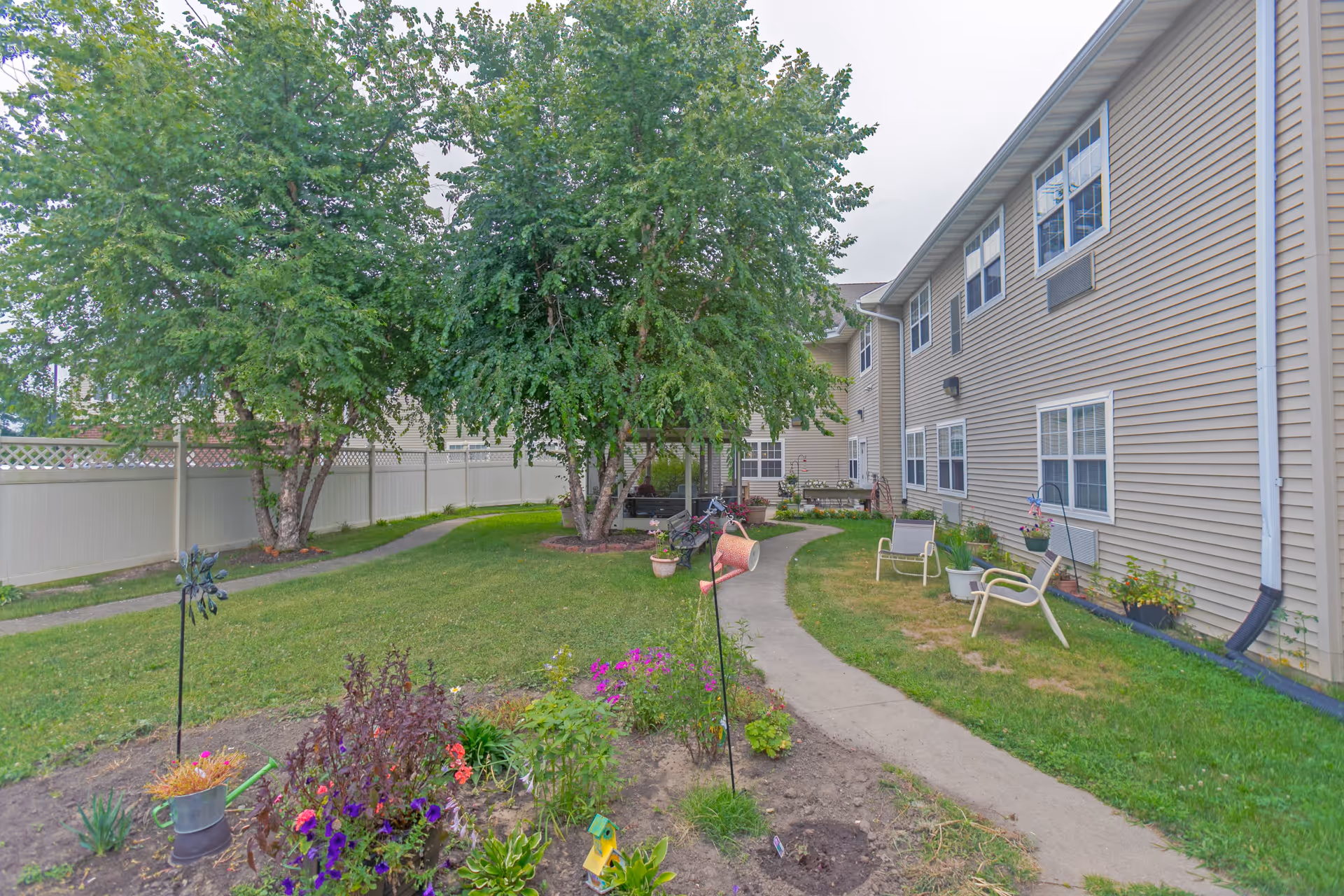 Outdoor garden area at Heritage Woods of Watseka featuring a curved concrete pathway, green grass, flower beds with colorful flowers, several trees, and beige residential building with multiple windows. There are a few chairs and potted plants along the pathway.