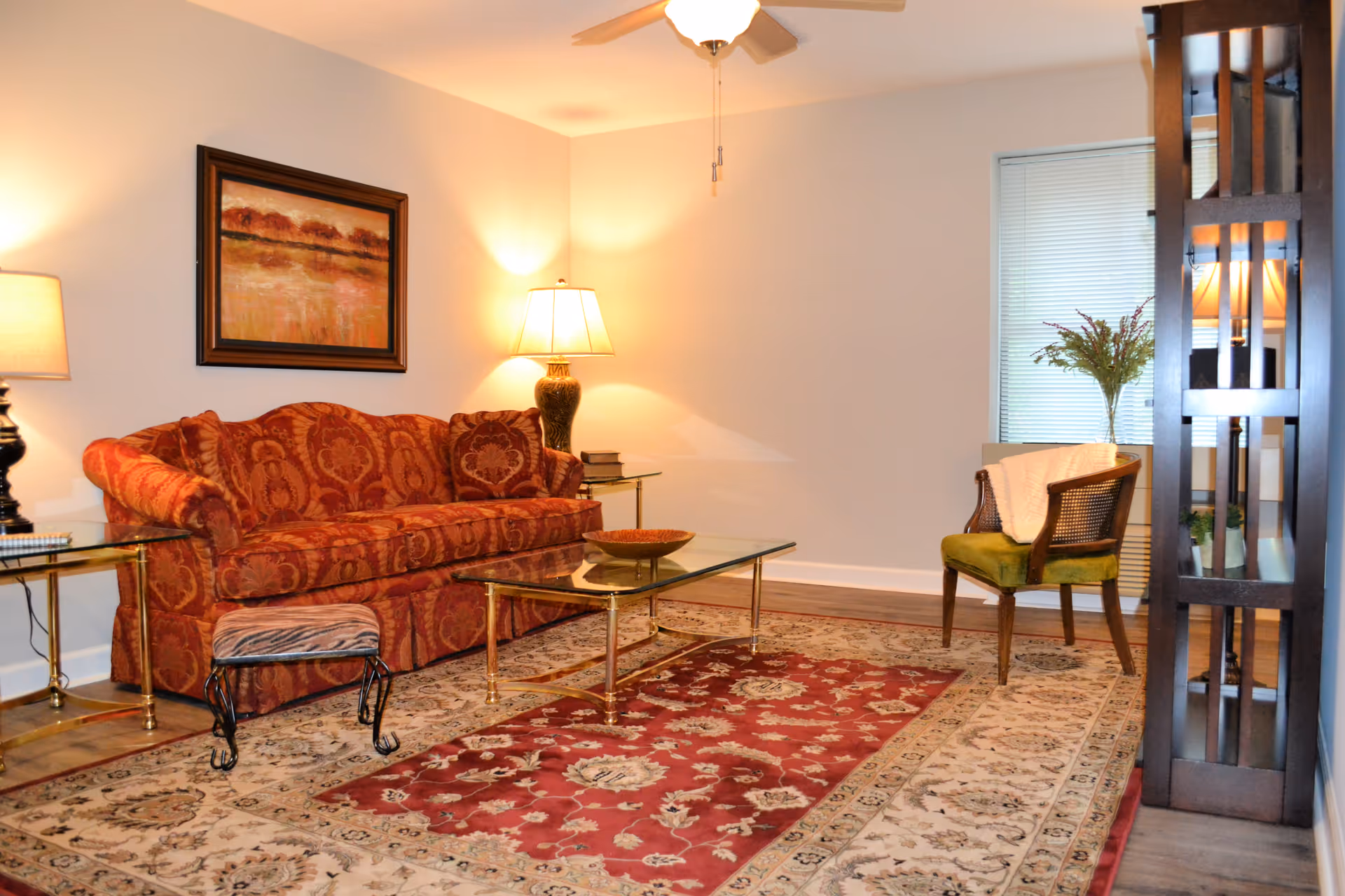 A cozy living room with a patterned red sofa, a glass coffee table with a decorative bowl, a small upholstered footstool, an armchair with a white throw blanket, two table lamps, a ceiling fan, a large area rug with red and beige floral patterns, a framed landscape painting on the wall, and a tall wooden shelving unit with plants and a lamp.