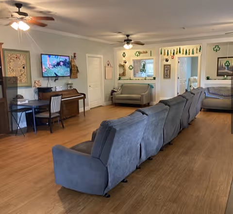 A spacious living room area with a row of gray recliner chairs facing a wall-mounted TV. The room has wooden flooring, ceiling fans with lights, a piano with a chair, and a small desk. There are two gray sofas along the walls, decorative wall hangings, and green shamrock decorations around a window and doorways.