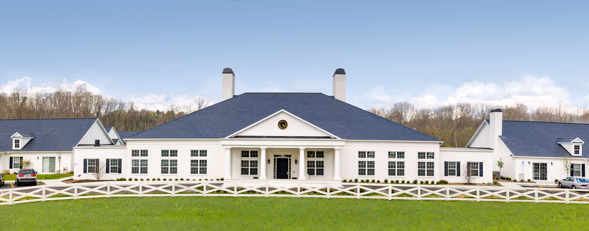 Front exterior view of Middleton Senior Living facility featuring a large white building with a dark blue roof, multiple windows, two chimneys, a central entrance with columns, and a clock above the entrance. The building is surrounded by a white wooden fence and green grass, with trees and a partly cloudy sky in the background.