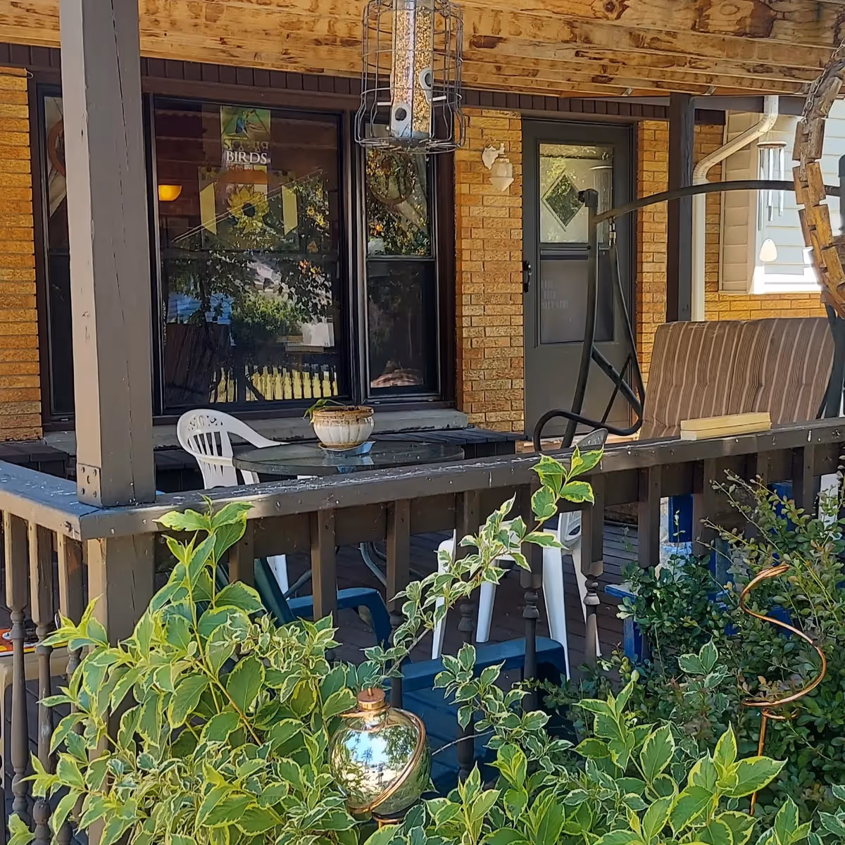 A covered porch area with a glass-top table, white plastic chairs, a cushioned swing, and various green plants in the foreground. The porch has wooden railings and a brick wall with windows and a door in the background. A bird feeder hangs from the porch ceiling.