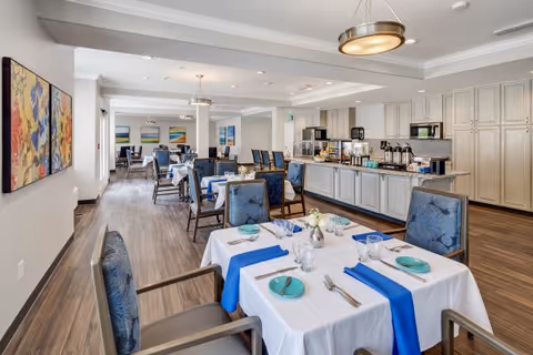 A bright and spacious dining room in a senior living facility with tables covered in white tablecloths and blue runners. Each table is set with plates, glasses, and silverware. Blue upholstered chairs surround the tables. The room has wooden flooring, modern light fixtures, and colorful artwork on the walls. A buffet area with coffee dispensers and cabinets is visible along one side of the room.