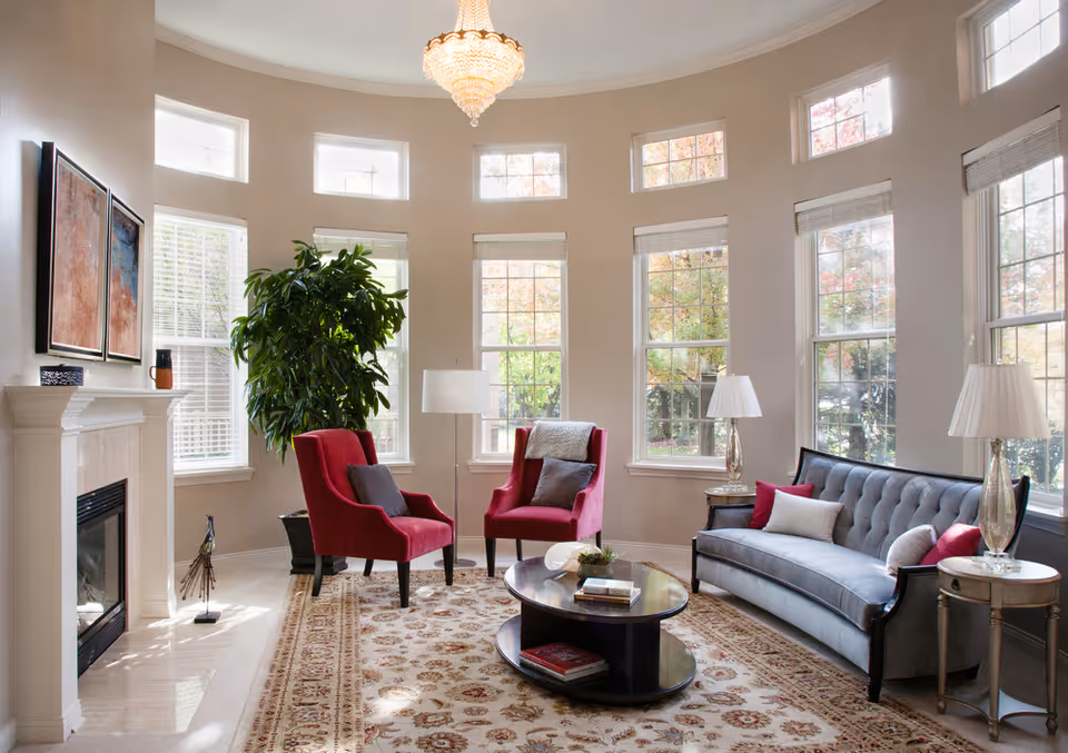 A bright living room with large windows letting in natural light. The room features two red armchairs with gray cushions, a gray tufted sofa with red and white pillows, a round dark wood coffee table with books and decorative items, two side tables with lamps, a large leafy plant, a fireplace with a white mantel, and two framed artworks above the fireplace. A patterned area rug covers the floor, and a chandelier hangs from the ceiling.
