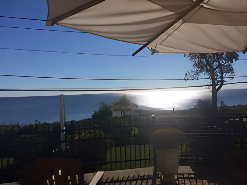 Patio view with an umbrella and railing overlooking the ocean and a sunlit shoreline.