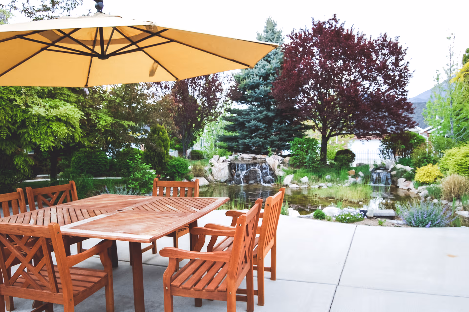 Outdoor patio area with a wooden table and chairs under a large beige umbrella, surrounded by lush greenery, trees, and a small pond with a waterfall feature in the background.