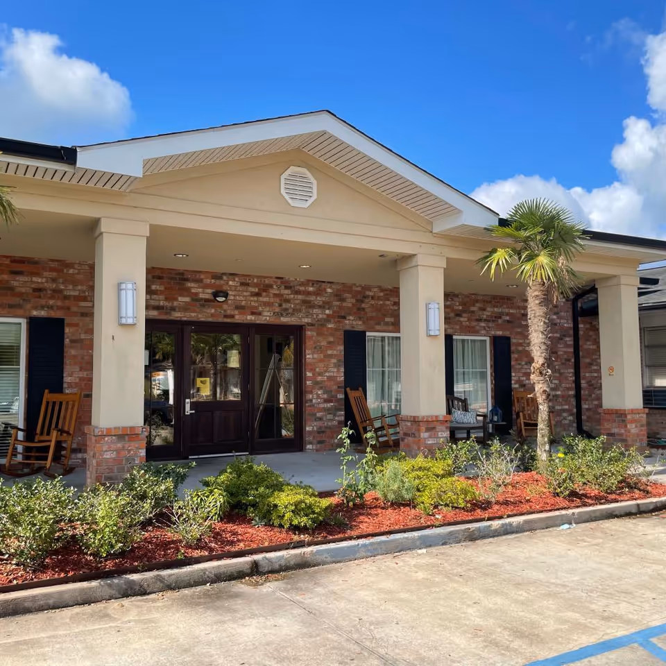 Front exterior view of Gonzales Healthcare Center showing a brick and beige building with a covered entrance, two columns, rocking chairs on the porch, landscaped bushes, a small palm tree, and a clear blue sky with some clouds.