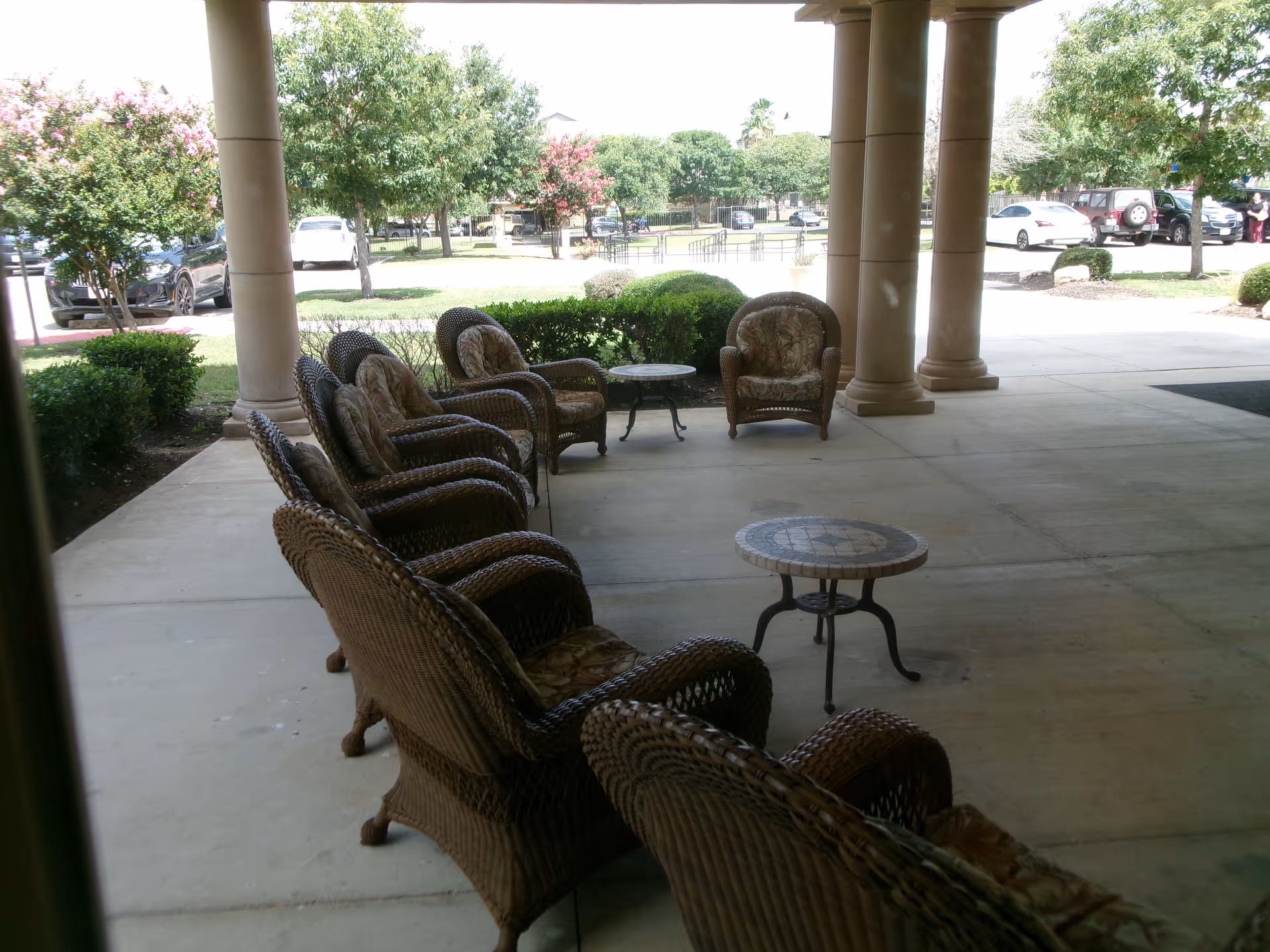 Outdoor covered patio area with several wicker chairs arranged around two small round tables. The patio overlooks a parking lot with cars and is surrounded by trees and bushes.