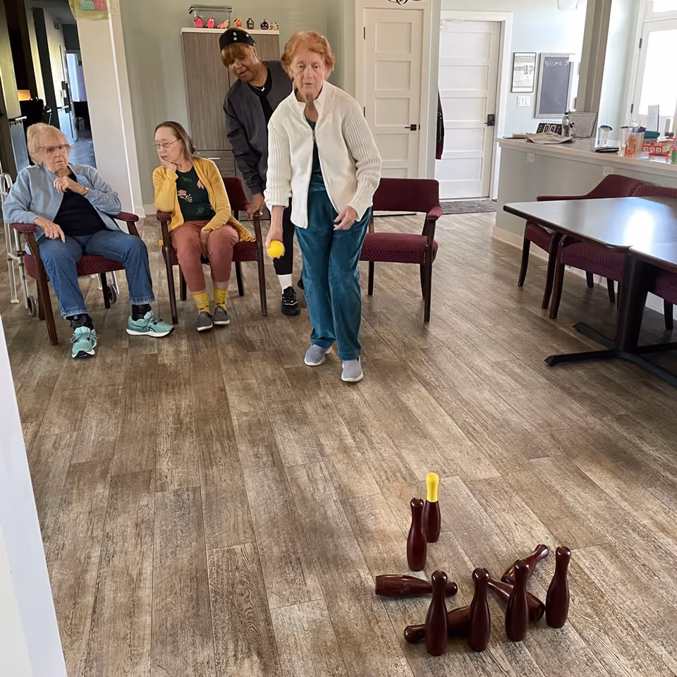 Four elderly women in a common room at a senior living facility. One woman is standing and rolling a yellow ball towards a set of small bowling pins on the floor, while the other three women are seated and watching. The room has wooden flooring, chairs, a table, and a cabinet in the background.