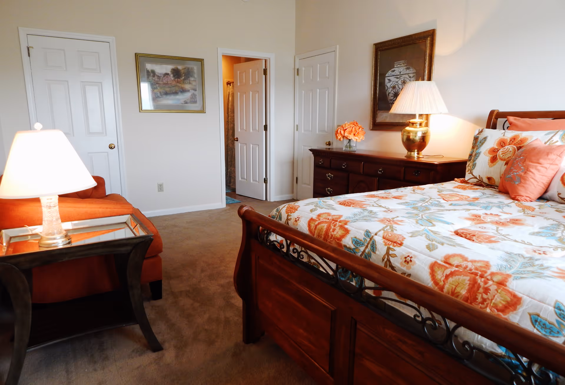 A cozy bedroom with a wooden bed frame featuring floral patterned bedding in shades of orange, blue, and white. There is a wooden dresser with a lamp and a vase of orange flowers on top. A framed picture hangs above the dresser. To the left, there is an orange armchair next to a side table with a lamp. Two doors are visible, one leading to a bathroom and the other likely a closet. The room has beige carpeting and light-colored walls.