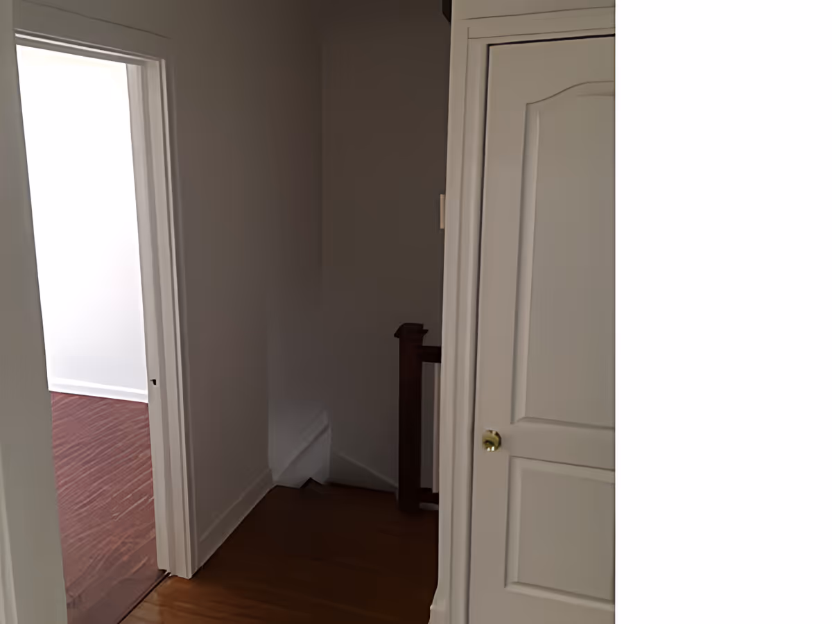 A hallway with wooden flooring leading to a room with dark wood flooring. There is a white door with a brass knob on the right side and a stair railing visible in the background.
