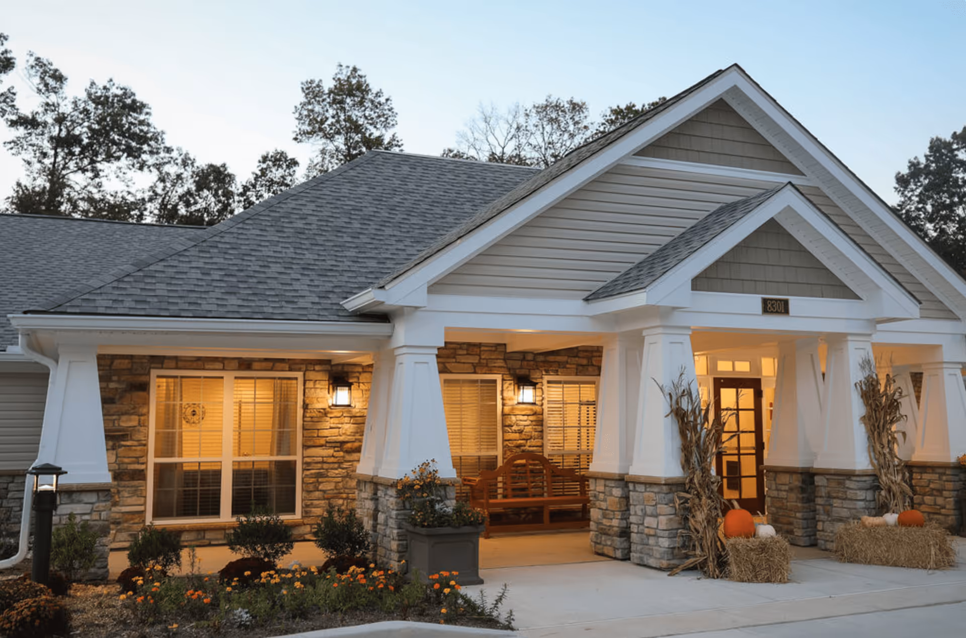 Exterior view of a senior living facility entrance with stone and siding facade, a covered porch with white columns, fall decorations including pumpkins and hay bales, and warm lighting inside.