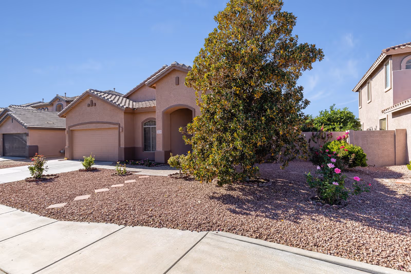 Exterior view of a single-story house with a tiled roof, a garage, and a front yard landscaped with gravel, a large tree, and flowering plants under a clear blue sky.