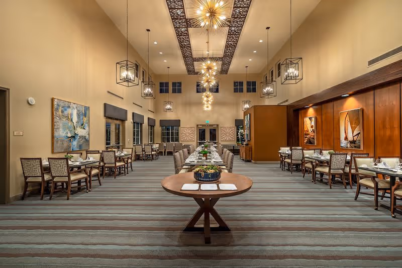 A spacious and well-lit dining room with long tables set for meals, surrounded by chairs. The room features high ceilings with decorative light fixtures, large windows with dark blinds, and artwork on the walls. A round table with a centerpiece is positioned in the foreground.