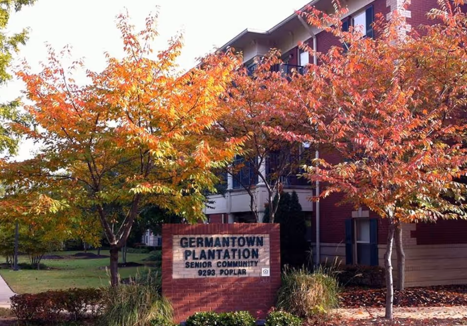 Exterior view of Germantown Plantation Senior Living Community building with autumn trees displaying orange and red leaves. A brick sign in front reads 'Germantown Plantation Senior Community 9293 Poplar'.