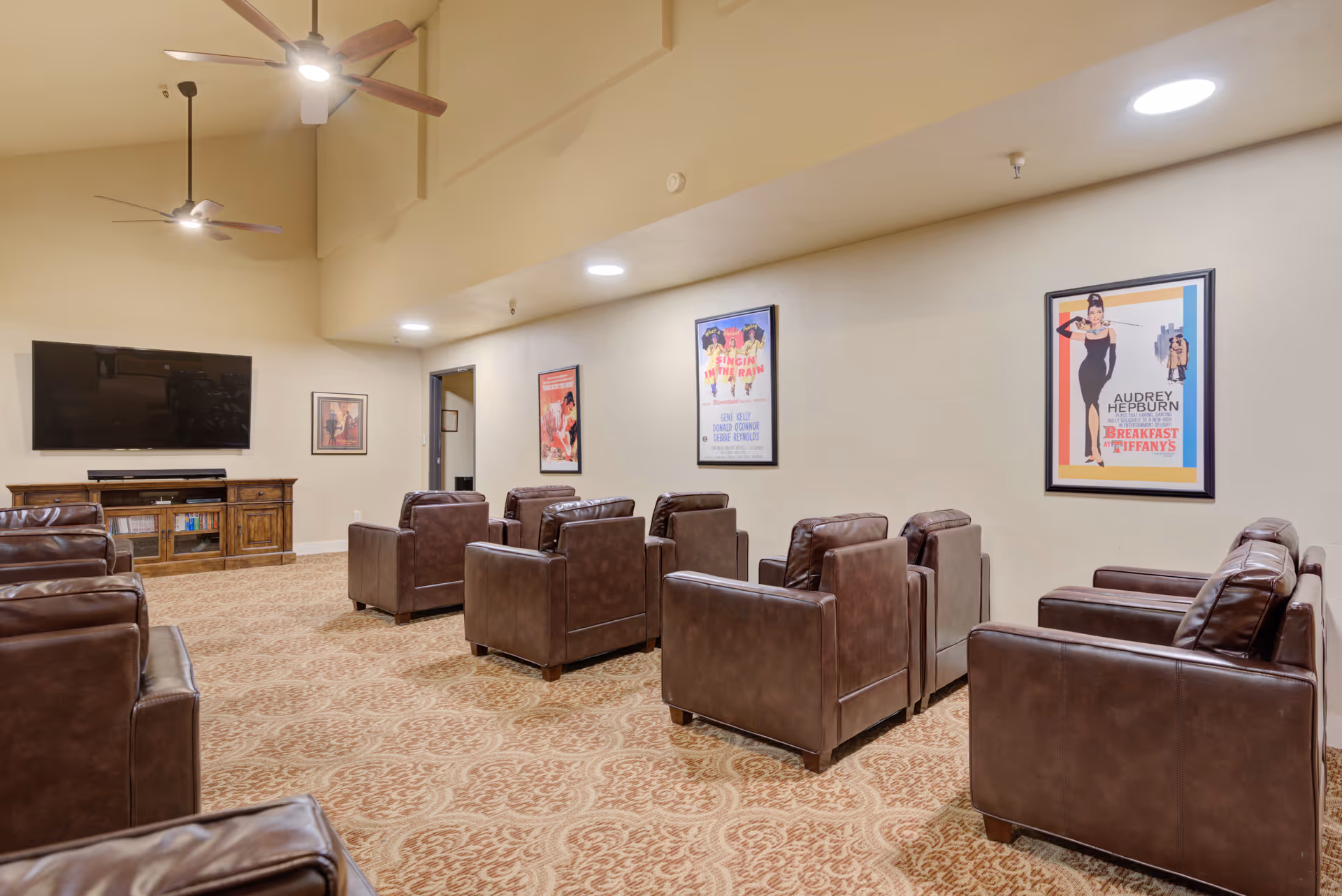 A cozy media room with multiple brown leather armchairs arranged in rows facing a large flat-screen TV mounted on a beige wall. The room has a patterned carpet, ceiling fans with lights, and framed vintage movie posters on the walls.