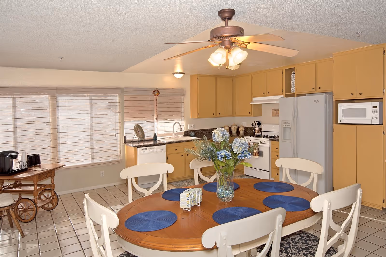 Bright kitchen with a round wooden dining table set with blue placemats, yellow cabinets, and a ceiling fan.