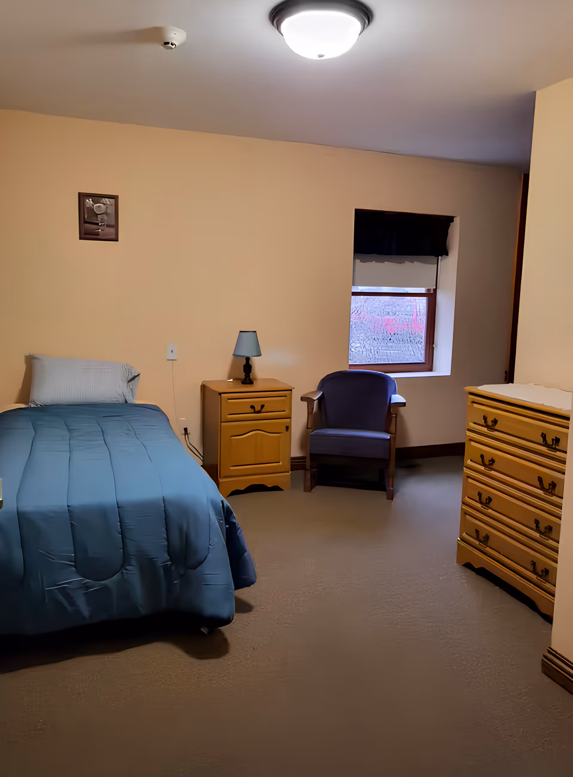 A tidy single bedroom with a blue bedspread, wooden nightstand and dresser, and an armchair by the window.