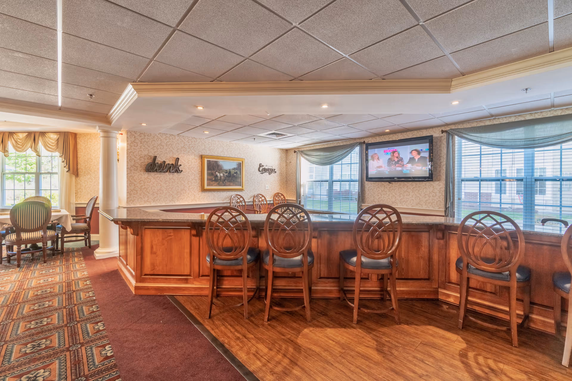 Wood-paneled bar with several high-backed stools in a comfortable dining/lounge area with windows, a mounted TV, and nearby dining tables.