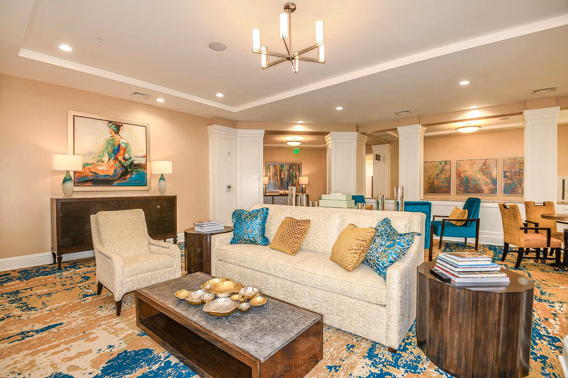 A well-lit living room area in a senior living facility featuring a beige sofa with blue and gold decorative pillows, a beige armchair, a wooden coffee table with decorative bowls, and a side table with stacked books. The room has beige walls, colorful abstract paintings, and a patterned carpet with blue and beige tones. There are also dining tables and chairs visible in the background.