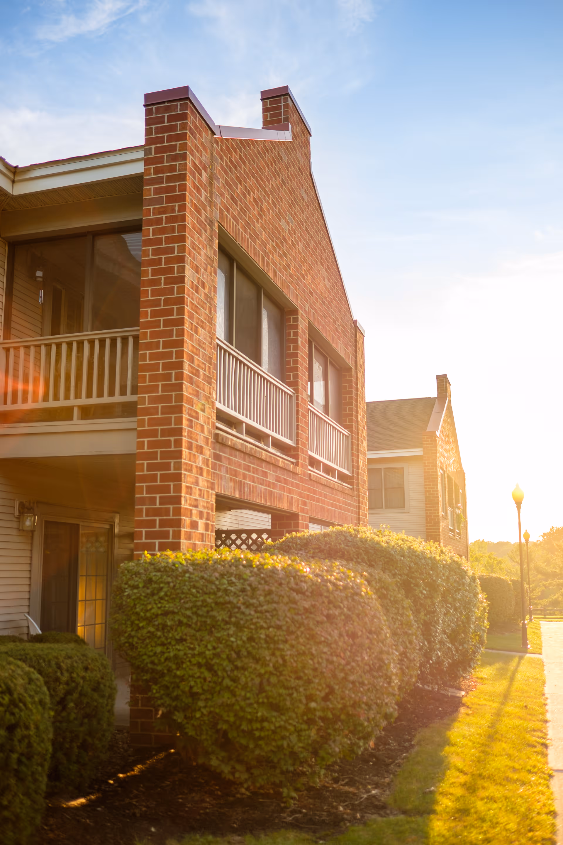 Exterior view of a brick building with balconies and neatly trimmed bushes in front, illuminated by warm sunlight during late afternoon or early evening.
