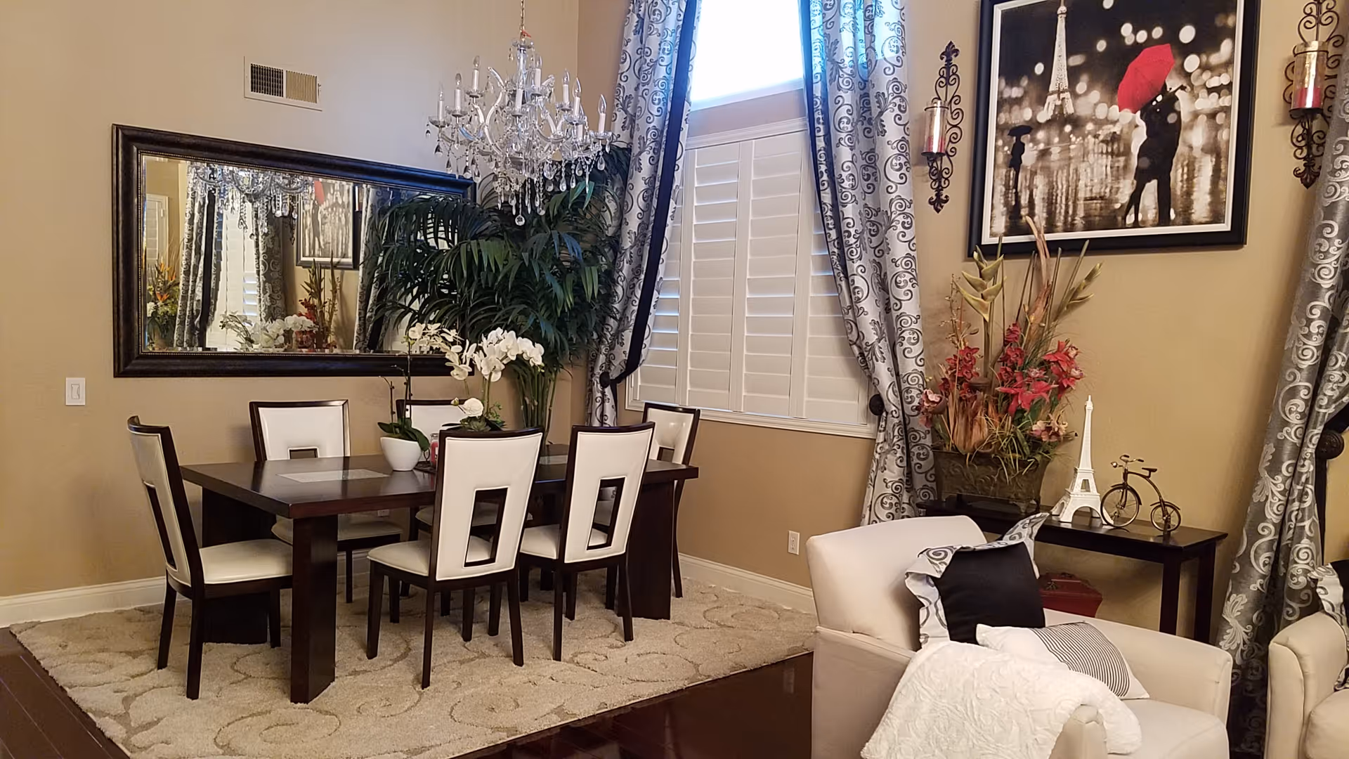 A dining room with a dark wooden table and six white cushioned chairs. A large mirror hangs on the beige wall above the table, reflecting a chandelier with crystal decorations. There are patterned curtains framing a window with white shutters. A large green plant and a floral arrangement sit near the window. On the right, a small table holds a miniature Eiffel Tower, a decorative bicycle, and a framed picture of a couple under a red umbrella in a black and white cityscape. A white armchair with pillows is partially visible in the foreground.