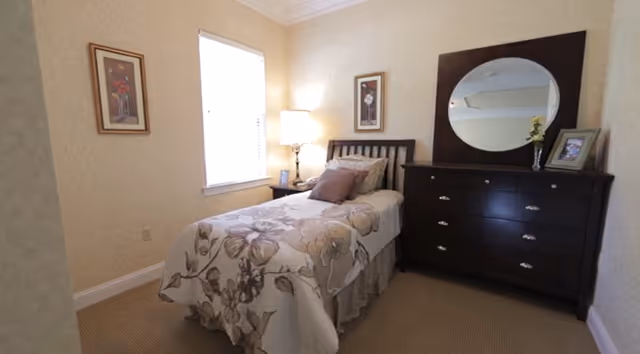 A small bedroom with a single bed covered in a floral bedspread. There is a wooden headboard, a bedside table with a lamp, and a dark wooden dresser with a round mirror above it. Two framed pictures hang on the walls, and a window with blinds lets in natural light.