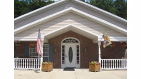 Front entrance of a single-story brick building with a white gabled porch, columns, flags, and planters.