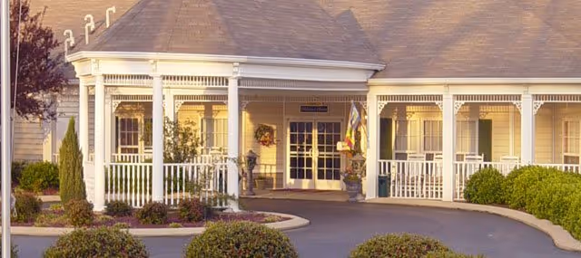 Front exterior view of Charter Senior Living of Paris building with a covered entrance, white columns, and a circular driveway surrounded by bushes and small trees.