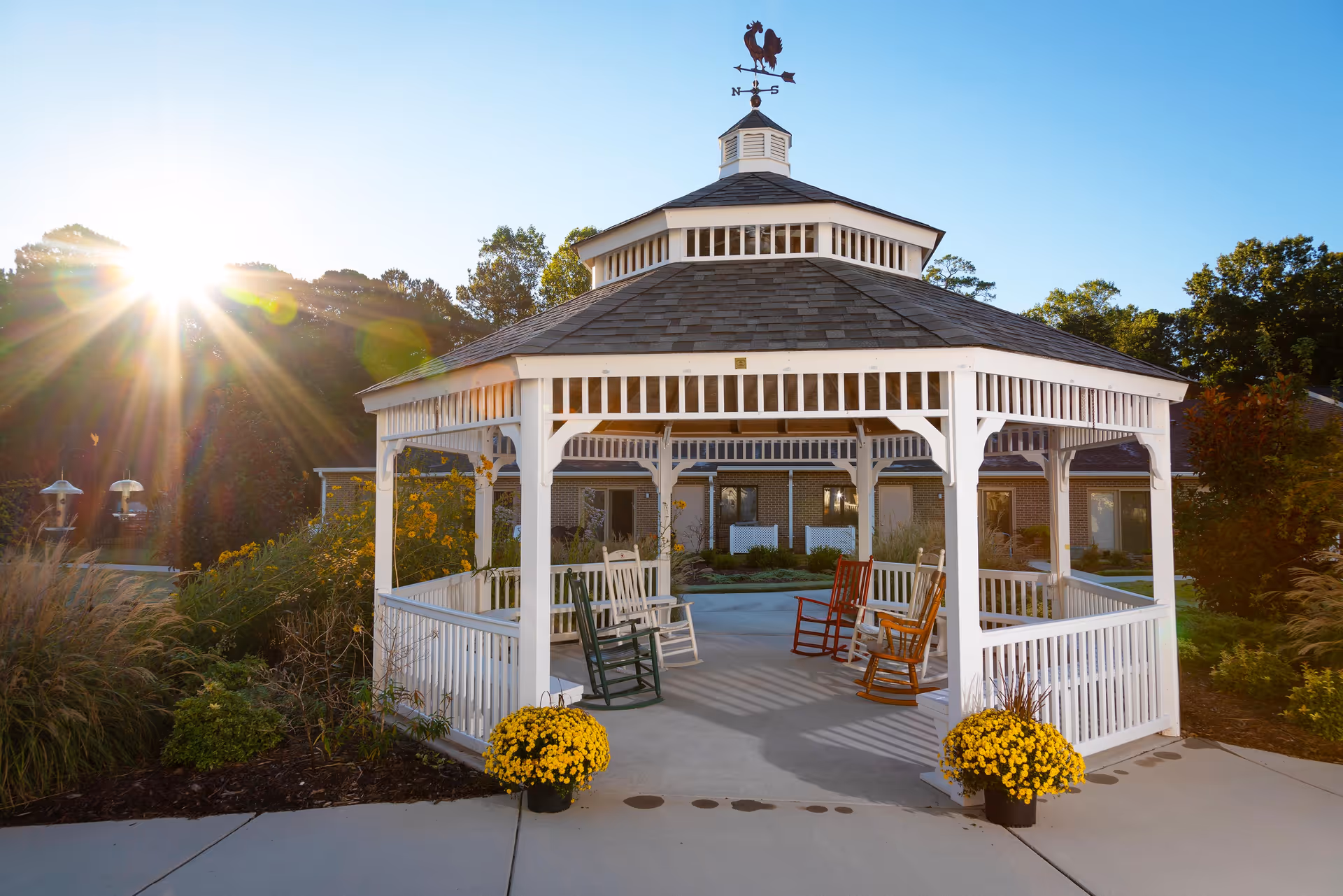 A white wooden gazebo with a weather vane on top, surrounded by greenery and flowers, with several colorful rocking chairs inside. The sun is shining brightly from the left side, casting long shadows on the concrete pathway around the gazebo.