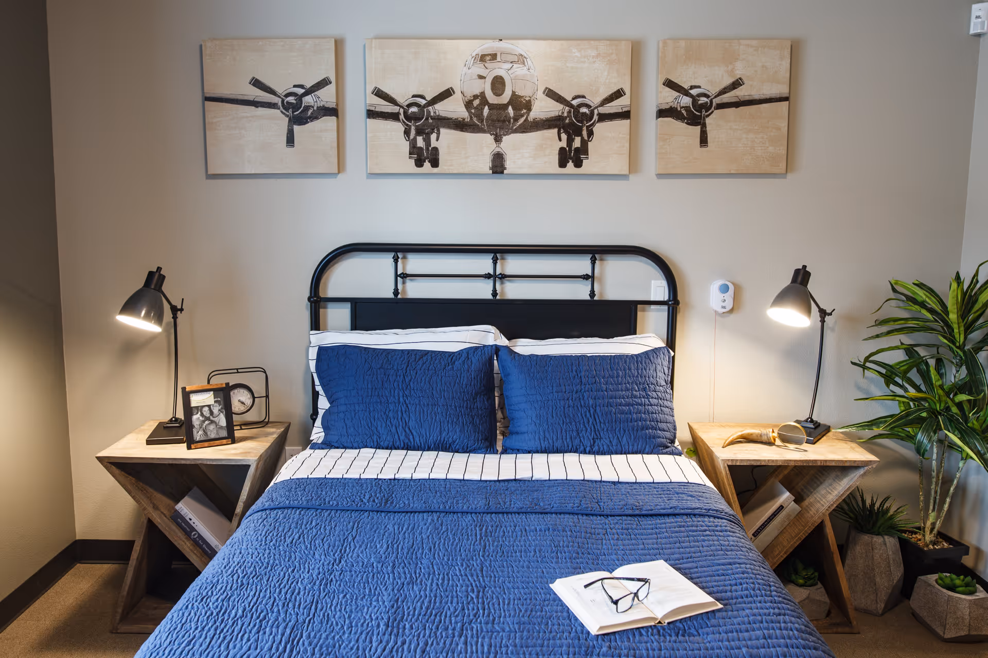 A neatly made bed with a black metal headboard, covered with a blue quilt and matching pillows. On the bed is an open book with a pair of glasses resting on it. On either side of the bed are wooden nightstands with modern black lamps, books, a framed photo, and decorative items. Above the bed is a three-panel artwork depicting the front view of an airplane. A green potted plant is placed on the right side of the bed against a beige wall.