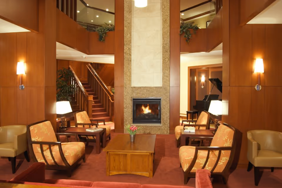 A cozy living room area with wooden panel walls, a central fireplace with a fire burning, and comfortable seating including armchairs with floral upholstery and beige chairs. There are side tables with lamps and books, a wooden coffee table with a small plant, and a grand piano in the background near a staircase.