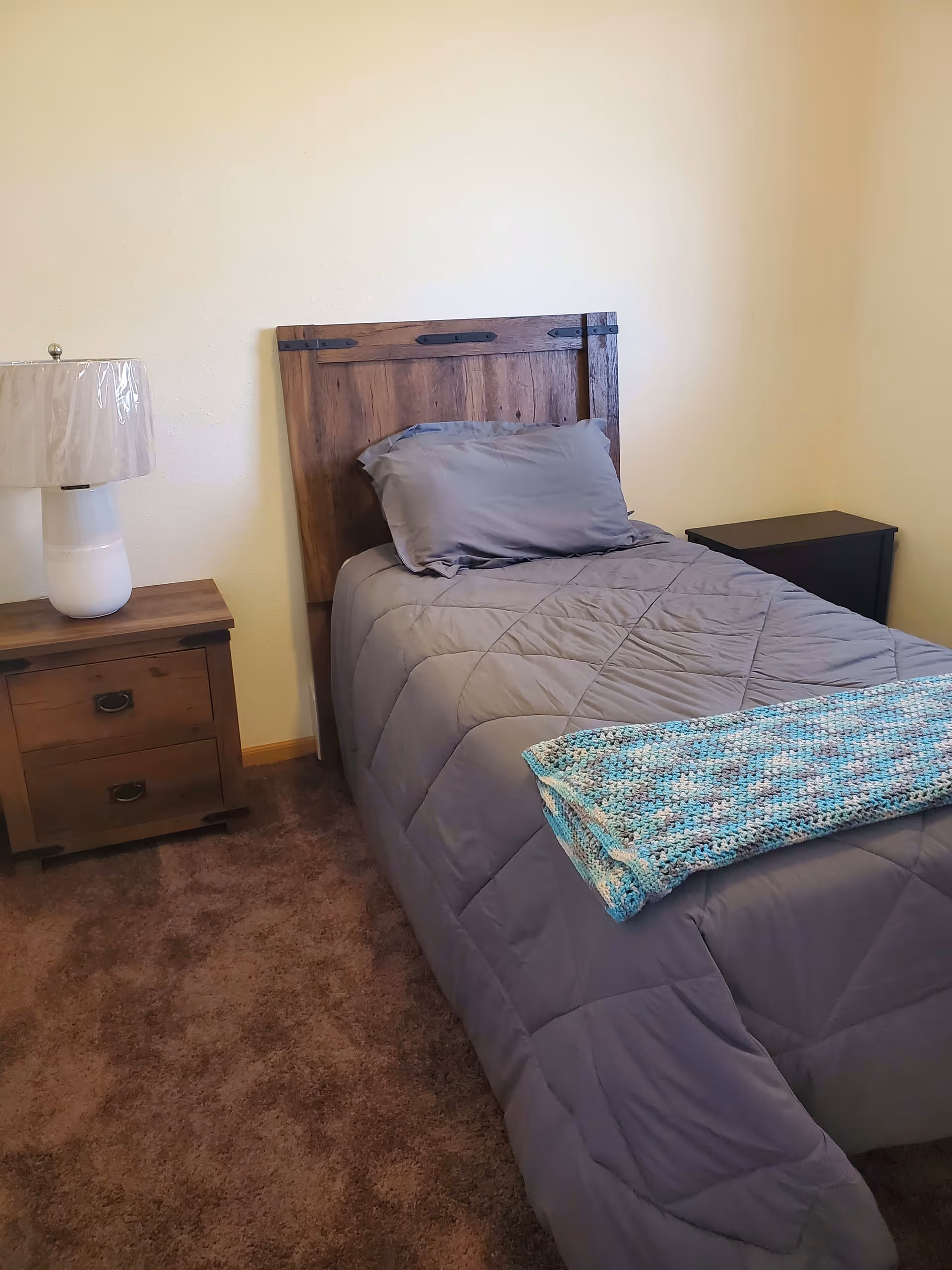 A small bedroom with a single bed featuring a wooden headboard, gray bedding, and a folded blue and white knitted blanket at the foot. To the left of the bed is a wooden nightstand with two drawers and a white lamp with a plastic cover on the shade. The floor is carpeted in brown, and the walls are painted a light beige color.