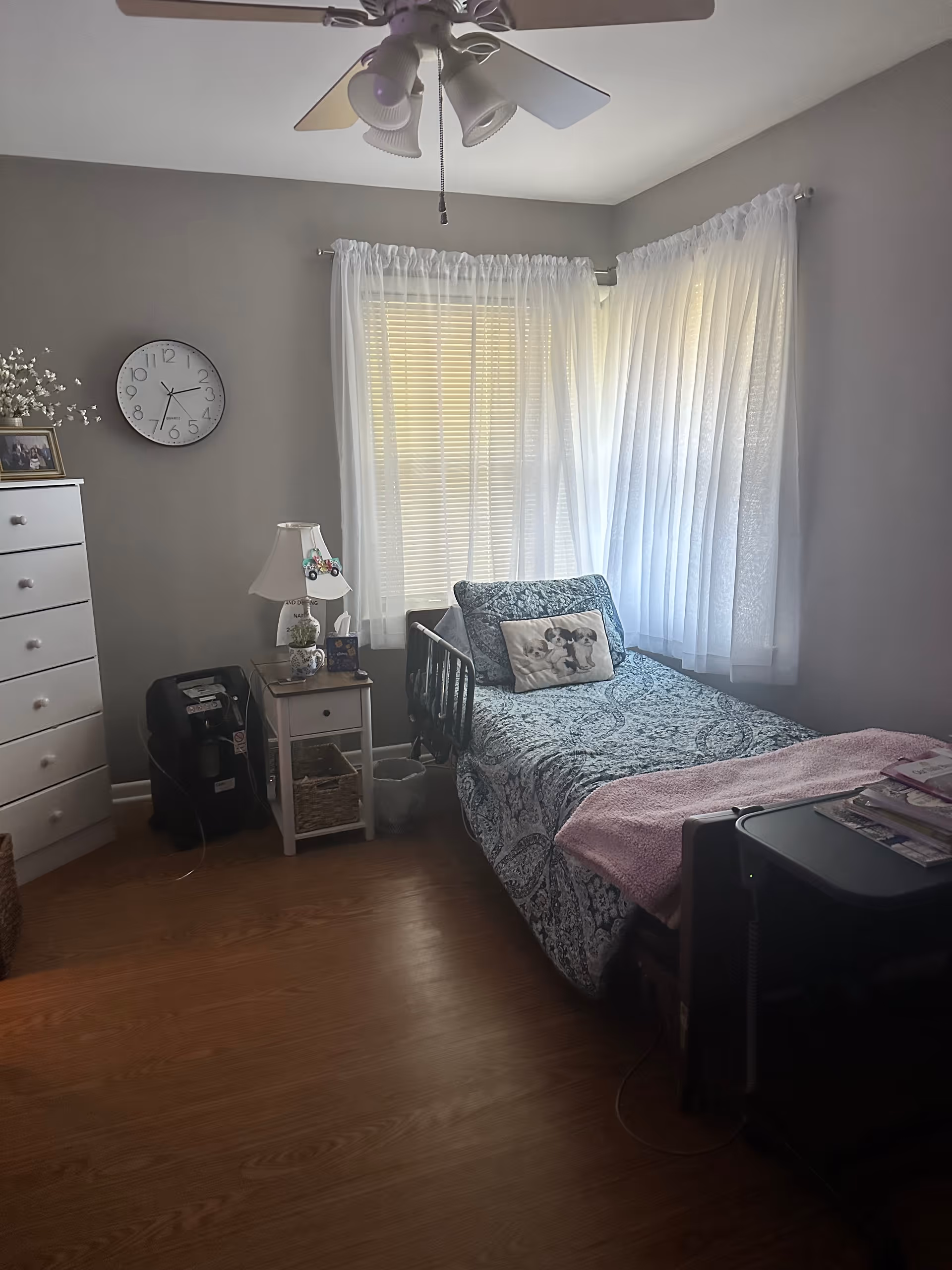 A small bedroom with a single bed covered in a blue patterned bedspread and a pink blanket at the foot. There is a pillow with a dog design on the bed. Next to the bed is a small white nightstand with a lamp, tissue box, and decorative items. A white dresser with drawers is on the left side of the room, and a wall clock hangs above it. The room has two windows with white sheer curtains allowing natural light to enter. The floor is wooden, and a ceiling fan with lights is visible on the ceiling.