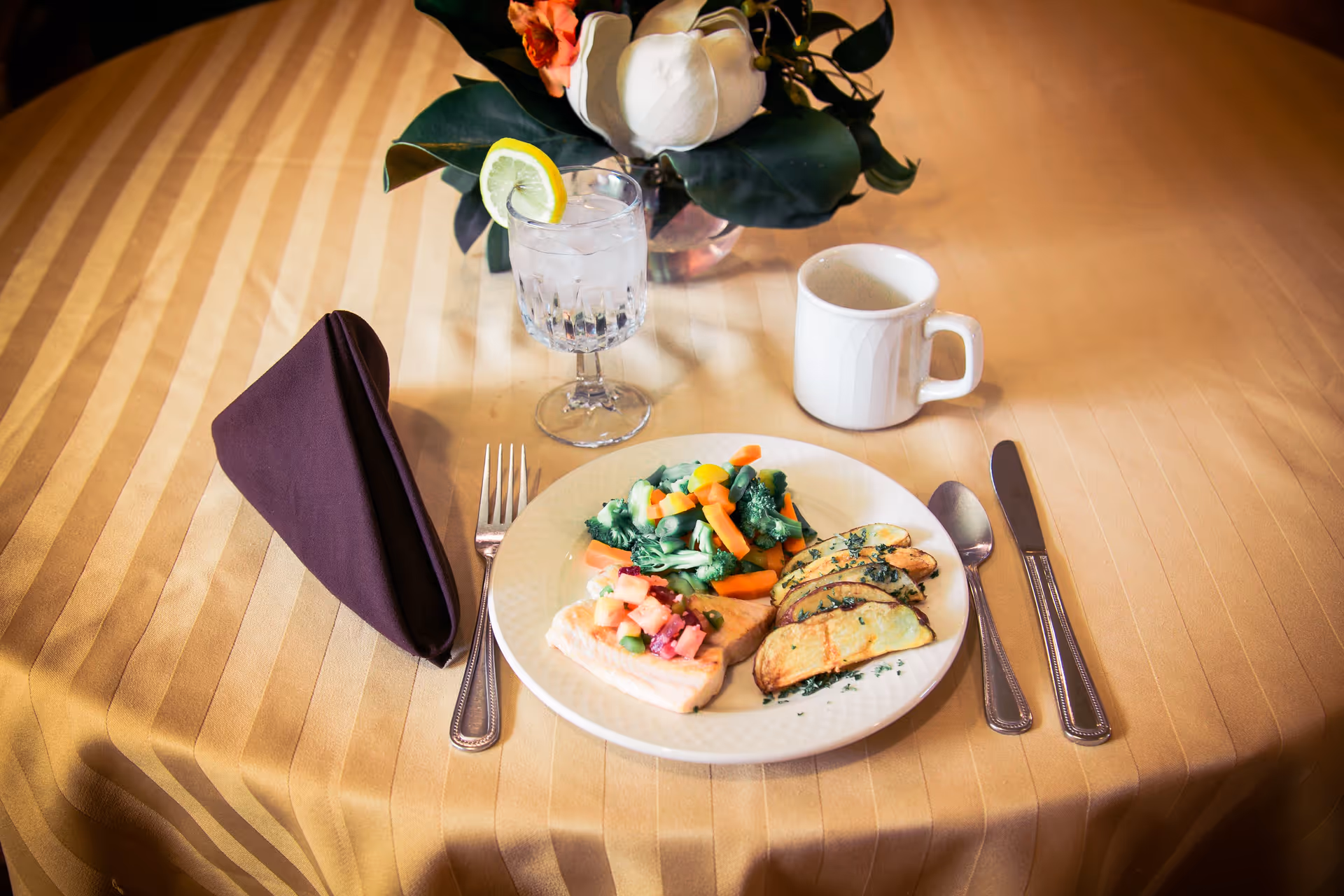 A neatly set dining table with a plate of food including grilled chicken topped with salsa, steamed mixed vegetables, and roasted potato wedges. The table has a beige striped tablecloth, a folded dark brown napkin, a fork, knife, and spoon. There is a glass of water with a lemon slice and a white coffee mug. A floral centerpiece with white and orange flowers is also on the table.
