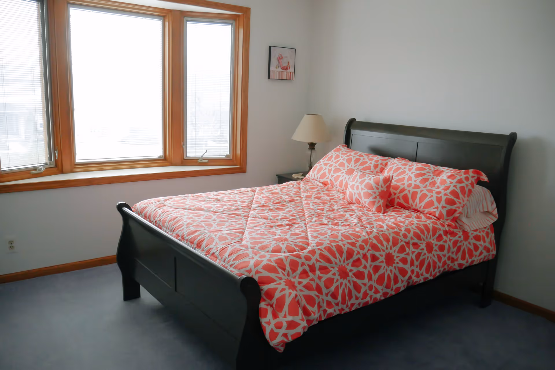 A bedroom with a dark wooden bed frame and a bed made with a red and white patterned comforter and pillows. There is a window with wooden trim and blinds on the left wall, a small framed picture on the wall above a nightstand with a lamp.