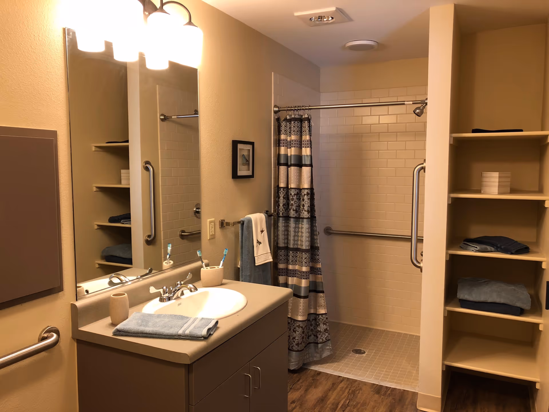 A bathroom with a sink and countertop on the left, a large mirror above the sink, and a walk-in shower with a patterned shower curtain on the right. There are grab bars near the sink and inside the shower. Shelves with folded towels and a box are visible on the right side. The floor has a wood-like finish.