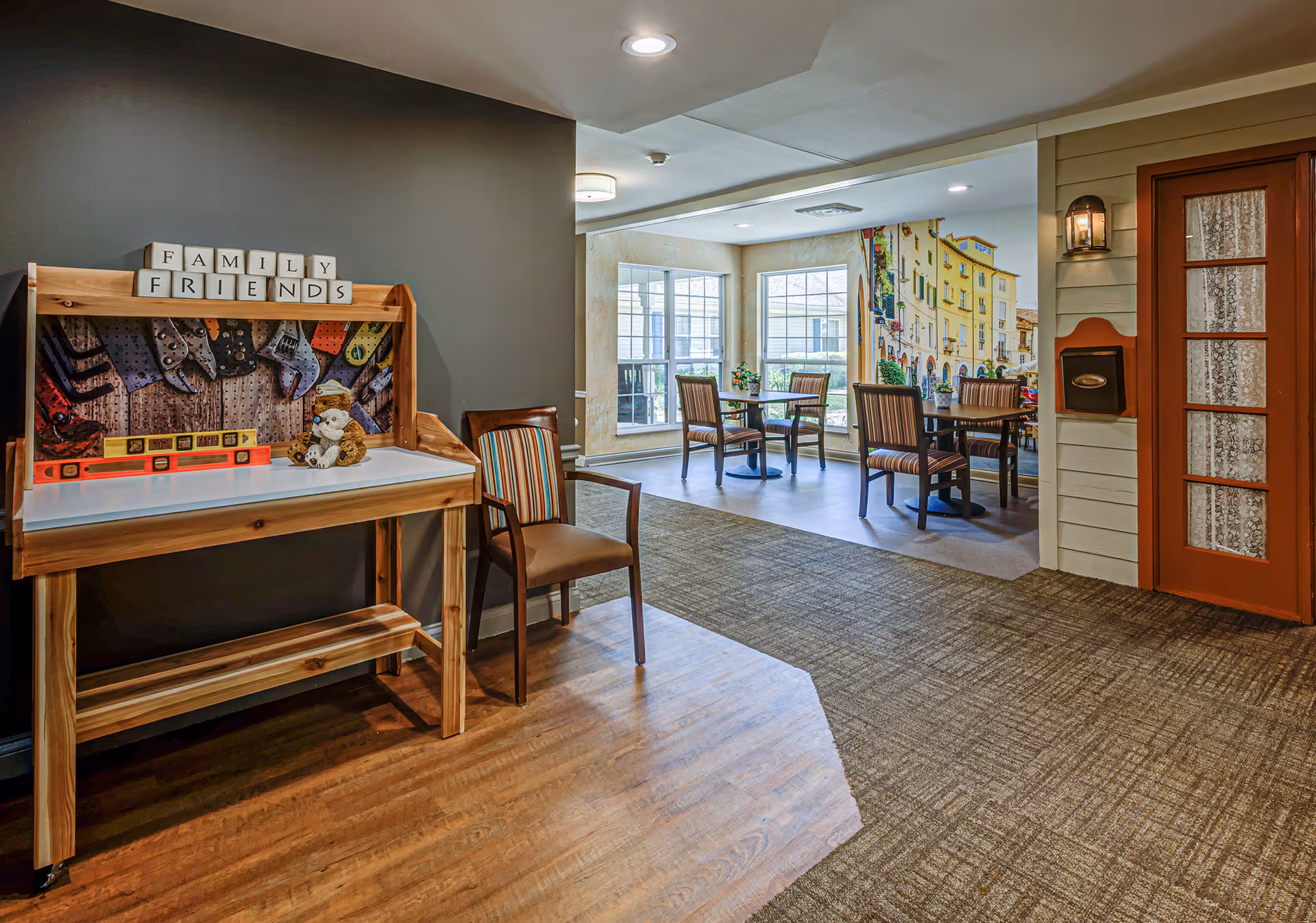 Interior view of a senior living facility common area with a wooden activity table on the left holding a stuffed animal and blocks spelling FAMILY FRIENDS. There are two chairs nearby, and in the background, a dining area with tables and chairs is visible next to large windows and a wall mural depicting a colorful street scene. A door with glass panels and a wall-mounted mailbox are on the right.
