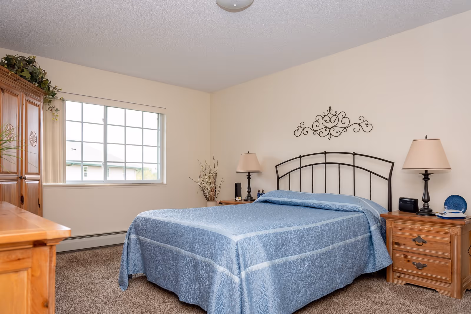 A bedroom with a large window, a bed with a blue quilted bedspread, two wooden nightstands with lamps, a wooden dresser, and a decorative metal wall art above the bed.