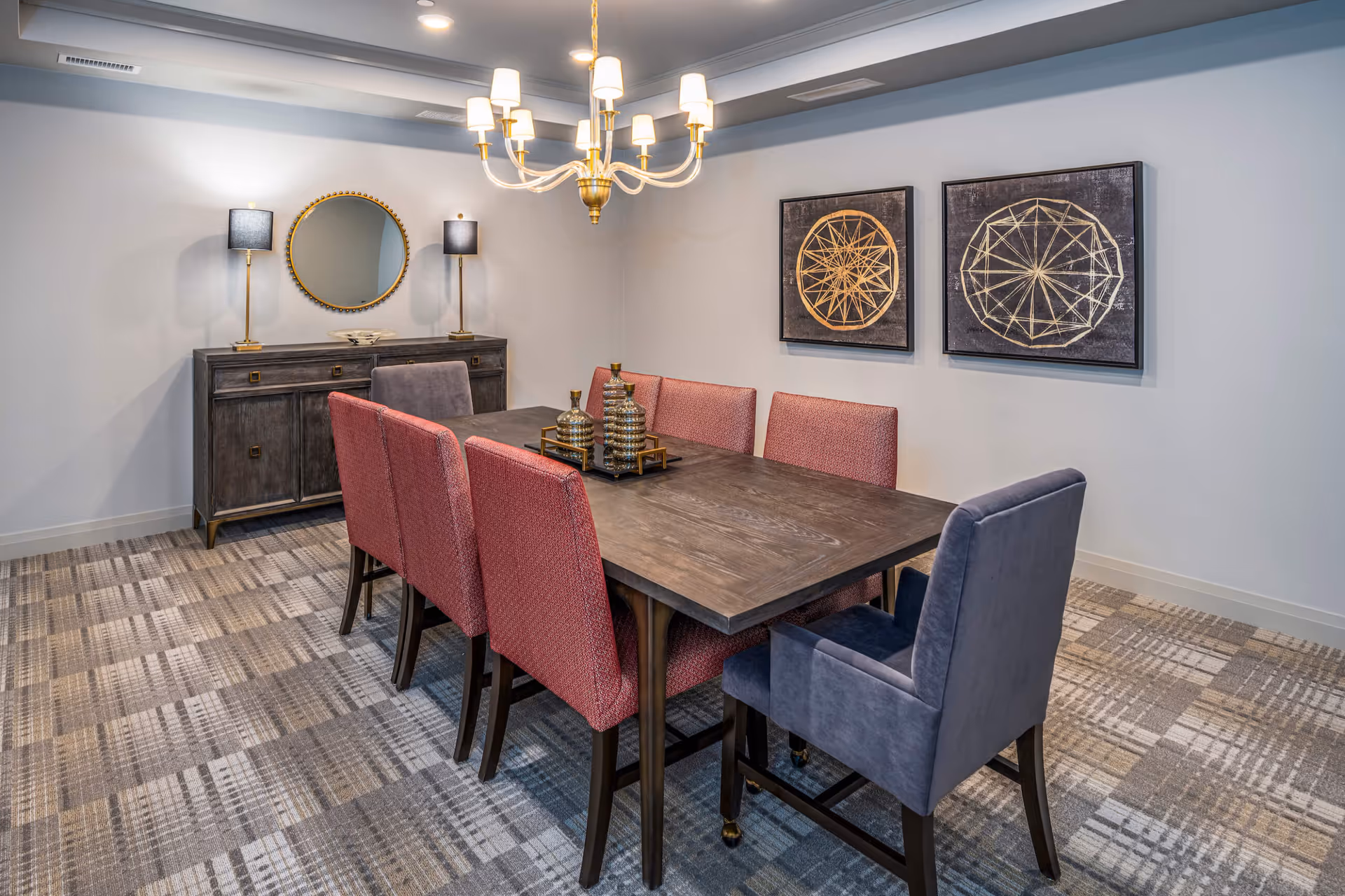A modern dining room with a rectangular wooden table surrounded by eight upholstered chairs, six in red and two in blue. Above the table hangs a gold chandelier with multiple lights. On the wall behind the table are two framed geometric art pieces. Against the far wall is a dark wooden sideboard with two black lamps and a round mirror above it. The floor is covered with a patterned carpet in neutral tones.