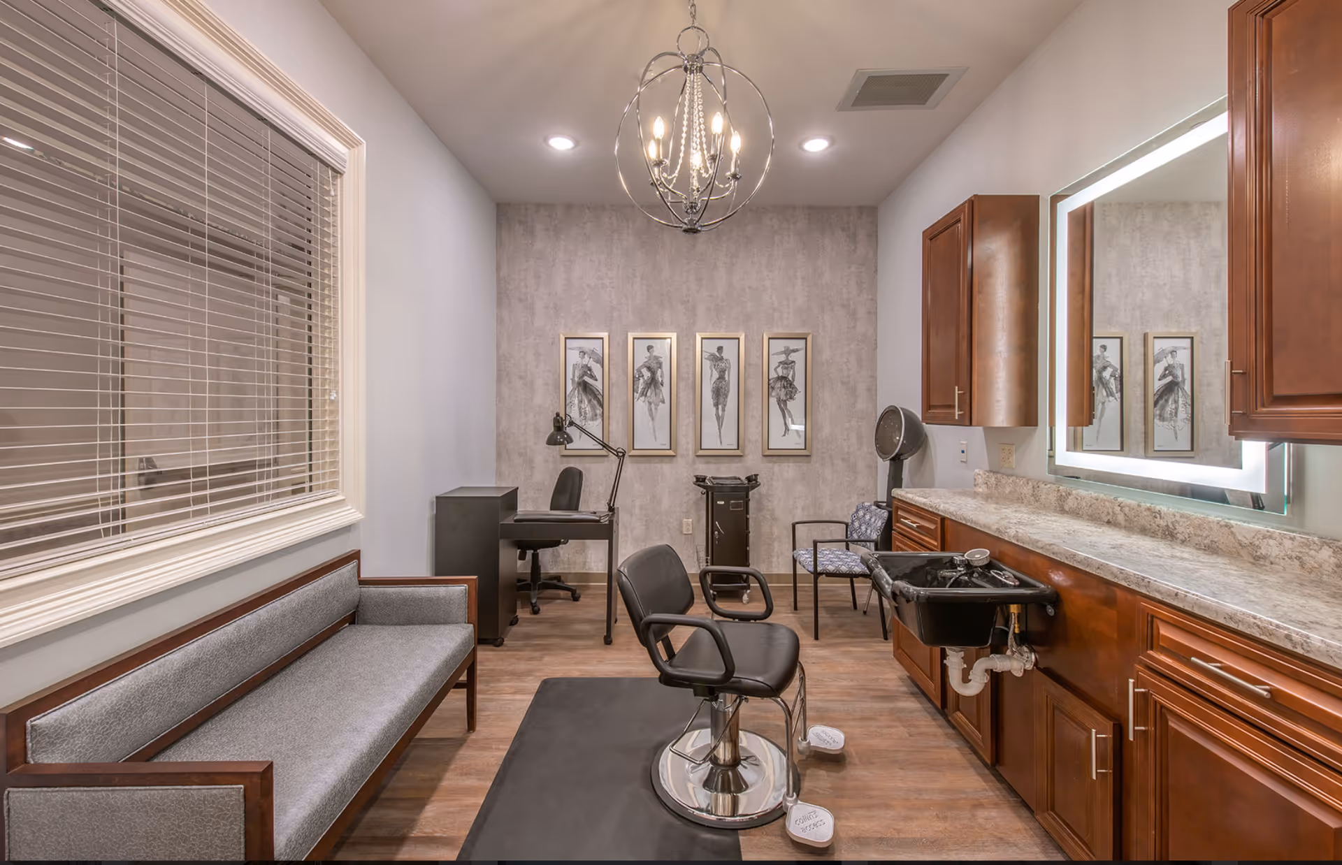 Interior view of a salon room with a black salon chair on a black mat, a black sink for hair washing, wooden cabinets with a marble countertop, a large illuminated mirror, a gray cushioned bench, a desk with a black chair and lamp, and four framed fashion sketches on the back wall. The room has wood flooring and a modern chandelier hanging from the ceiling.