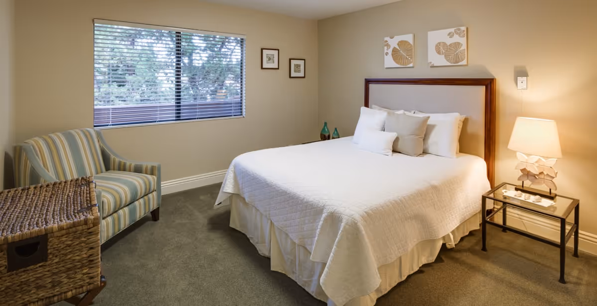 Well-lit furnished bedroom with a neatly made bed, bedside table and lamp, an armchair, window with blinds, and wall artwork.