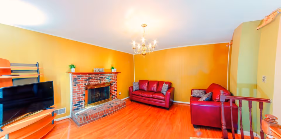 A cozy living room with yellow walls, a brick fireplace with a mantle decorated with small plants and a 'HOME' sign, two red leather sofas with patterned cushions, a wooden TV stand with a flat-screen TV, and a chandelier hanging from the ceiling.