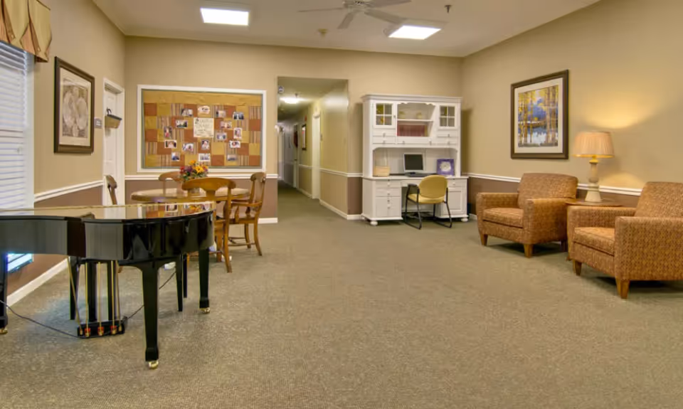 A senior living facility common area with a black grand piano on the left, a round wooden table with four chairs, a white desk with a computer and chair, two upholstered armchairs with a side table and lamp, and framed artwork on the beige walls.