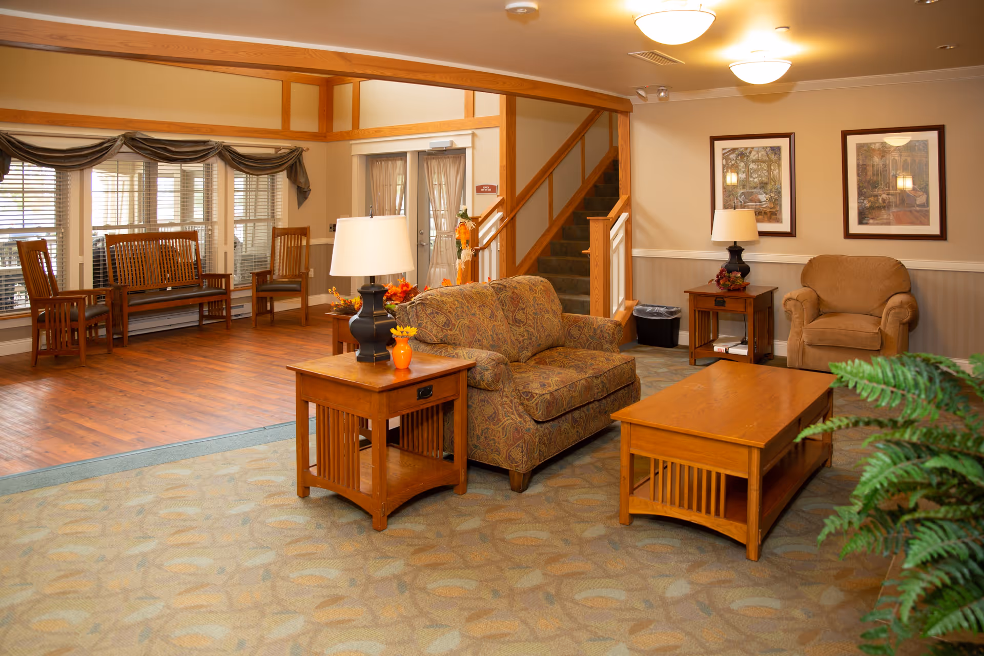 A cozy living room area in a senior living facility with a patterned loveseat, an armchair, wooden coffee and side tables, and lamps. There are framed pictures on the wall, a staircase leading upstairs, and a seating area with wooden chairs and a bench near large windows with blinds. The floor is a combination of carpet and wood, and there is a green plant in the foreground.