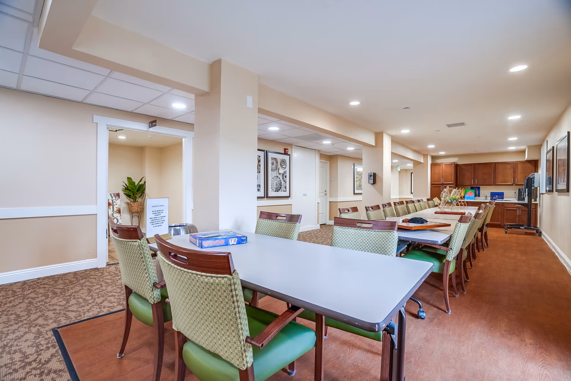 A long room with a large table surrounded by green cushioned chairs with wooden backs. The room has beige walls, recessed ceiling lights, and carpeted flooring. At the far end, there is a kitchenette area with wooden cabinets and a countertop. On the table, there are some board games and other items. The room appears to be a communal dining or activity space in a senior living facility.