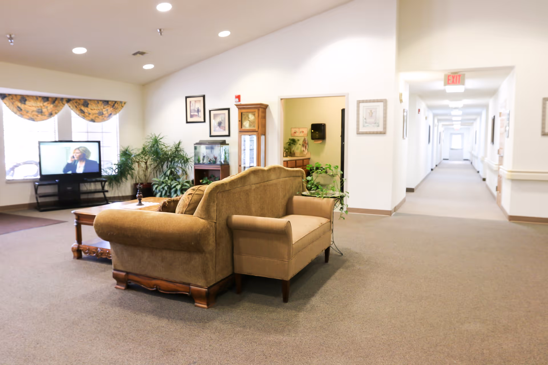 A cozy living room area in a senior living facility with a brown sofa and armchair facing a wooden coffee table. There is a television on a stand near windows with floral valances, several potted plants, framed pictures on the walls, and a long hallway extending to the right with an exit sign above a door at the end.