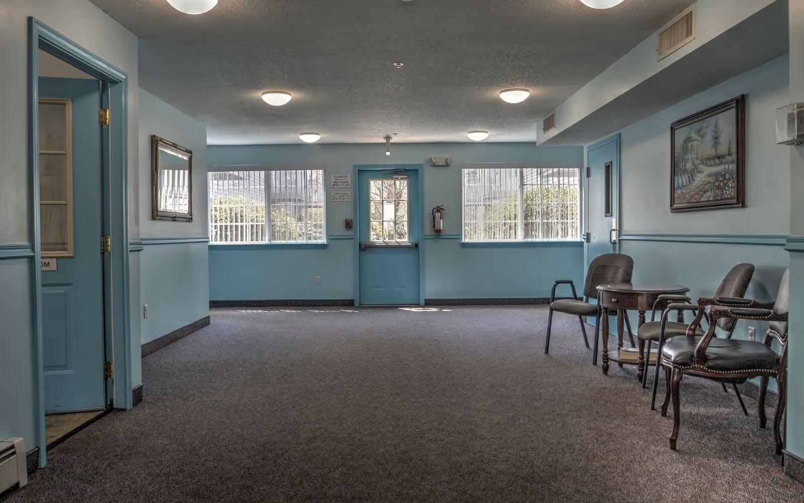 Light-blue painted common sitting area with chairs and small tables facing an exit door and windows.