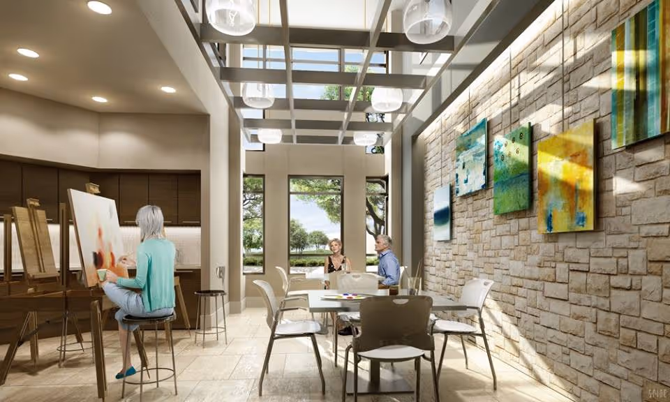 Bright senior living common room with residents painting and sitting at tables under a skylit ceiling and a stone wall hung with colorful artwork.